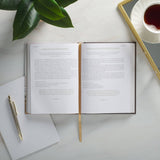 Open book on a white surface with a pen, plant, and cup of tea.