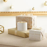 White candle on a beige surface with wrapped gifts and decorative lights in the background