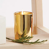 Gold candle holder with a lit candle and greenery on a wooden surface.