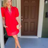 Woman in a red dress standing on a porch with a brown door.