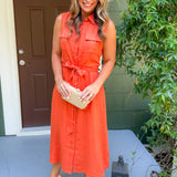 Woman in an orange dress standing on a porch with a brown door and greenery.