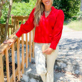 Woman in a red sweater and beige pants standing on a wooden deck with greenery in the background