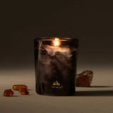 Lit candle in a decorative glass container with amber-colored stones on a dark background