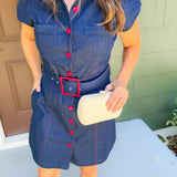 Woman wearing a denim dress with red buttons and a belt, holding a beige clutch.