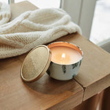 Candle in a decorative container on a wooden surface with a white textured blanket in the background