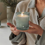 Person holding a lit Thymes candle in a cozy indoor setting