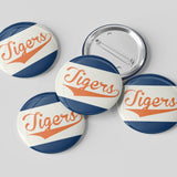 Five round pin-back buttons with a vintage sports design featuring the word 'Tigers' in orange script over diagonal navy and white stripes, shown on a light gray background with one button facing up and four facing down to show the metal pin-back mechanism.