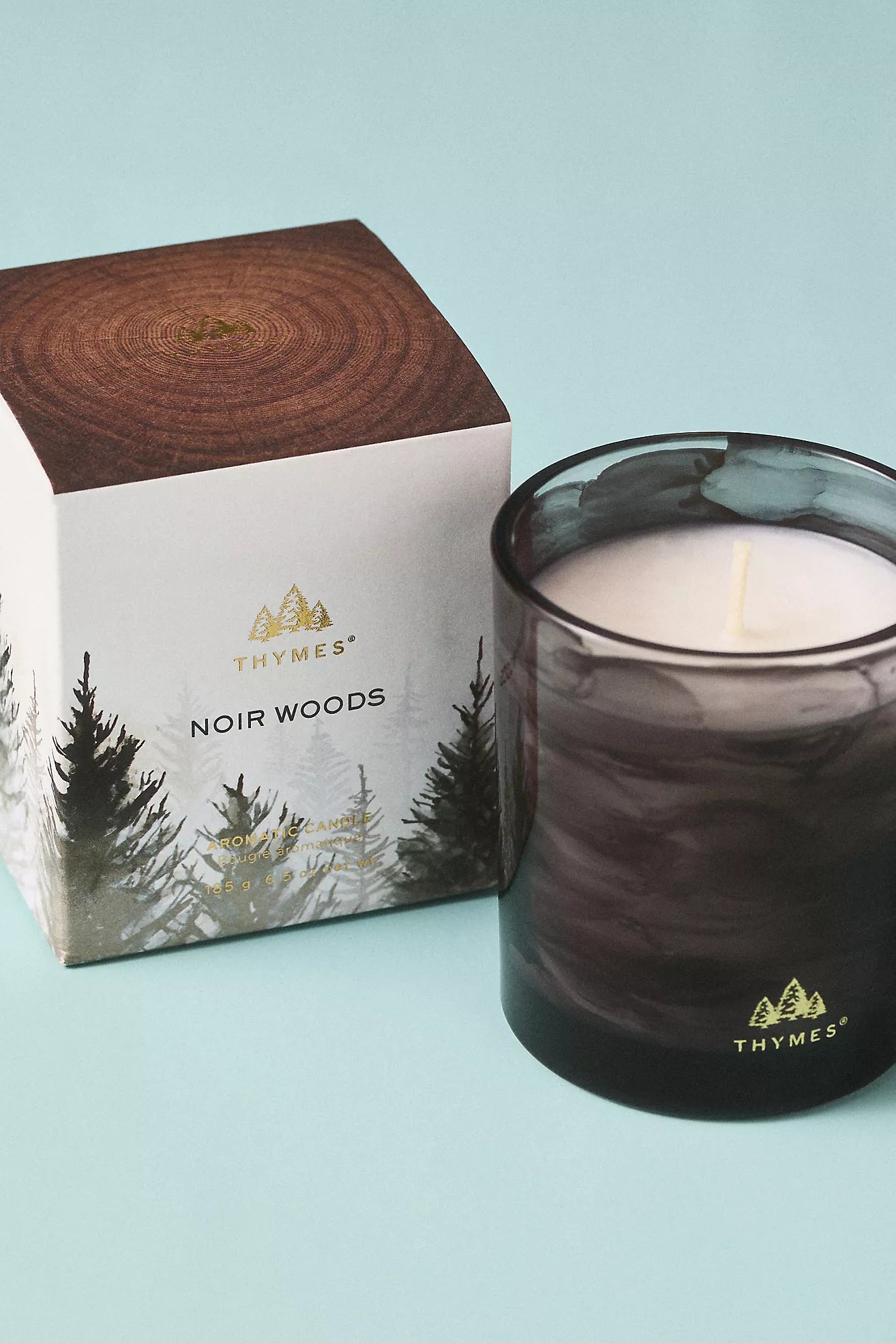 Thymes Noir Woods candle and packaging on a light blue background