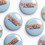 A repeating pattern of round light blue buttons with the text 'GAMEDAY in Auburn' printed in orange and dark blue, arranged on a white background.