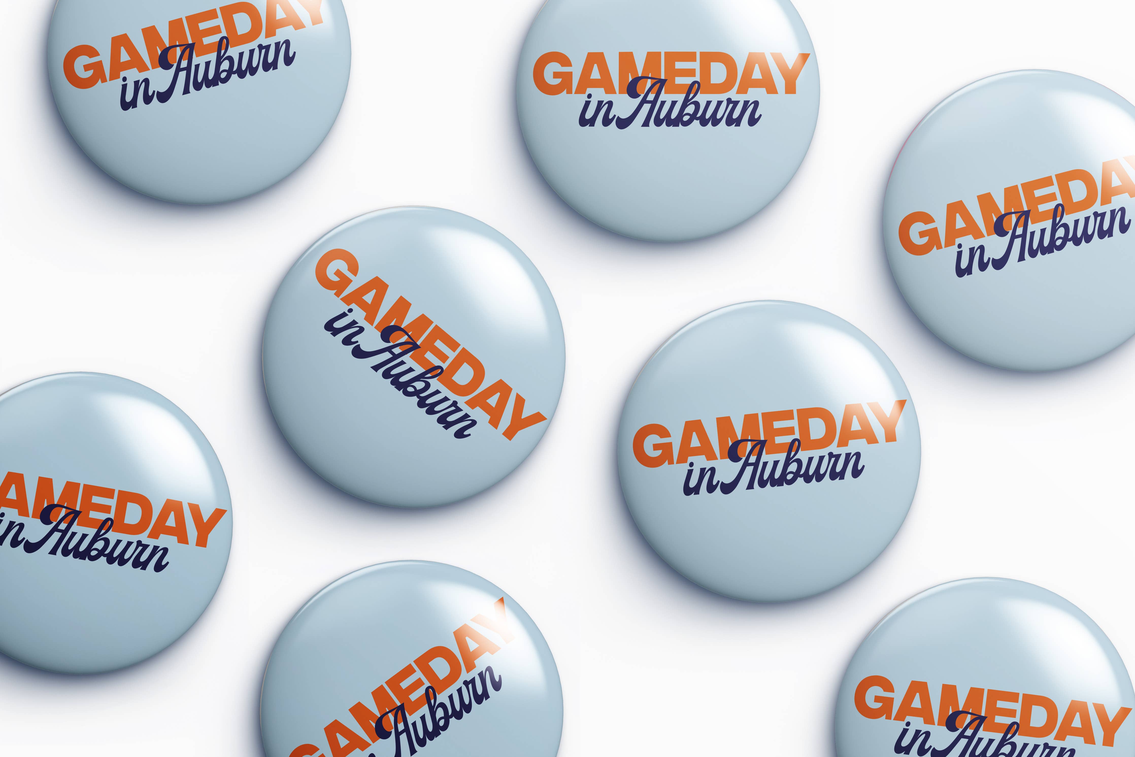 A repeating pattern of round light blue buttons with the text 'GAMEDAY in Auburn' printed in orange and dark blue, arranged on a white background.