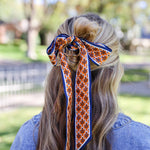 A woman viewed from behind wearing a patterned silk hair bow accessory in her hair, set against a blurred outdoor park background.