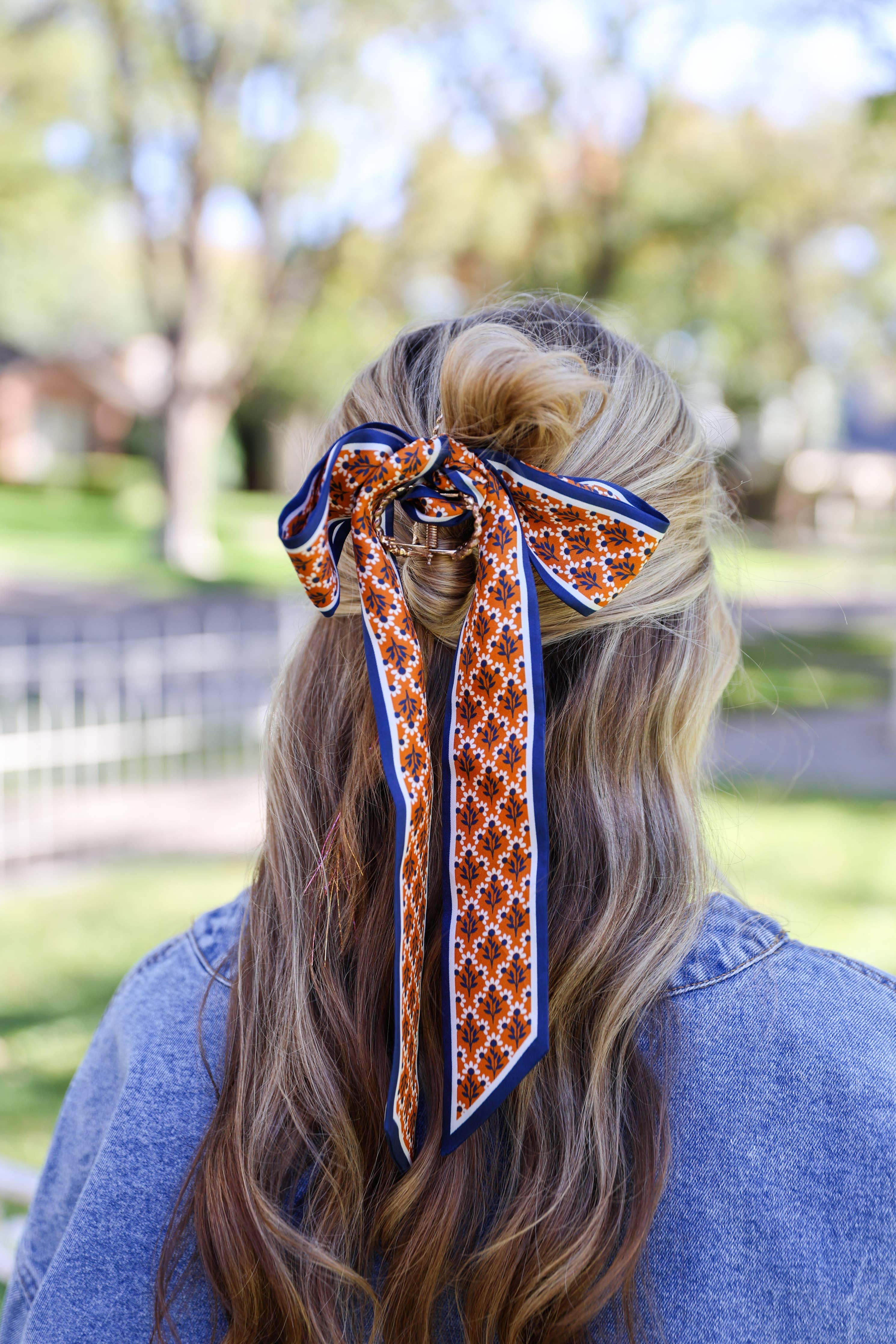 A woman viewed from behind wearing a patterned silk hair bow accessory in her hair, set against a blurred outdoor park background.
