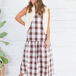 A woman modeling a brown and cream gingham midi dress with a cream striped vest, standing in front of a white wood paneled wall next to a potted plant.