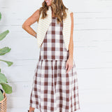A woman modeling a brown and cream gingham midi dress with a cream striped vest, standing in front of a white wood paneled wall next to a potted plant.