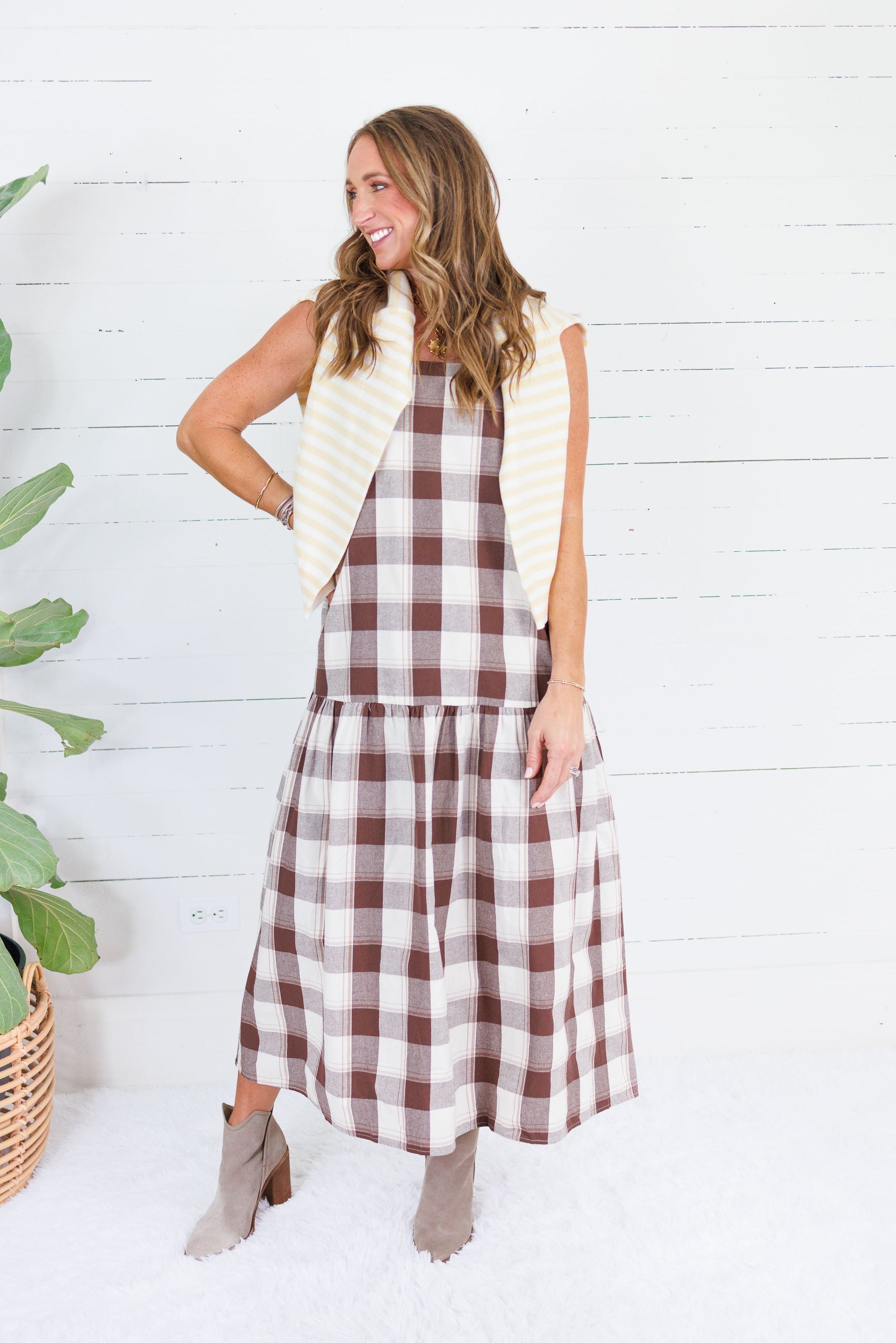 A woman modeling a brown and cream gingham midi dress with a cream striped vest, standing in front of a white wood paneled wall next to a potted plant.