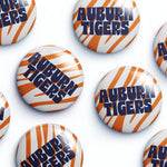 Several round button badges with the Auburn Tigers logo, featuring orange and white tiger stripes and navy blue lettering, arranged on a white background.