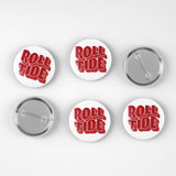Six round pin-back buttons on a light gray background, showing both the front and back views. The front features the words 'ROLL TIDE' in red 3D-style lettering on a white background, while the backs show a silver metal clasp.