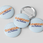 Five round pin-back buttons with a light blue background and the text 'GAMEDAY in Auburn' in orange and navy blue, with one button showing the silver metal pin-back mechanism, on a light gray background.