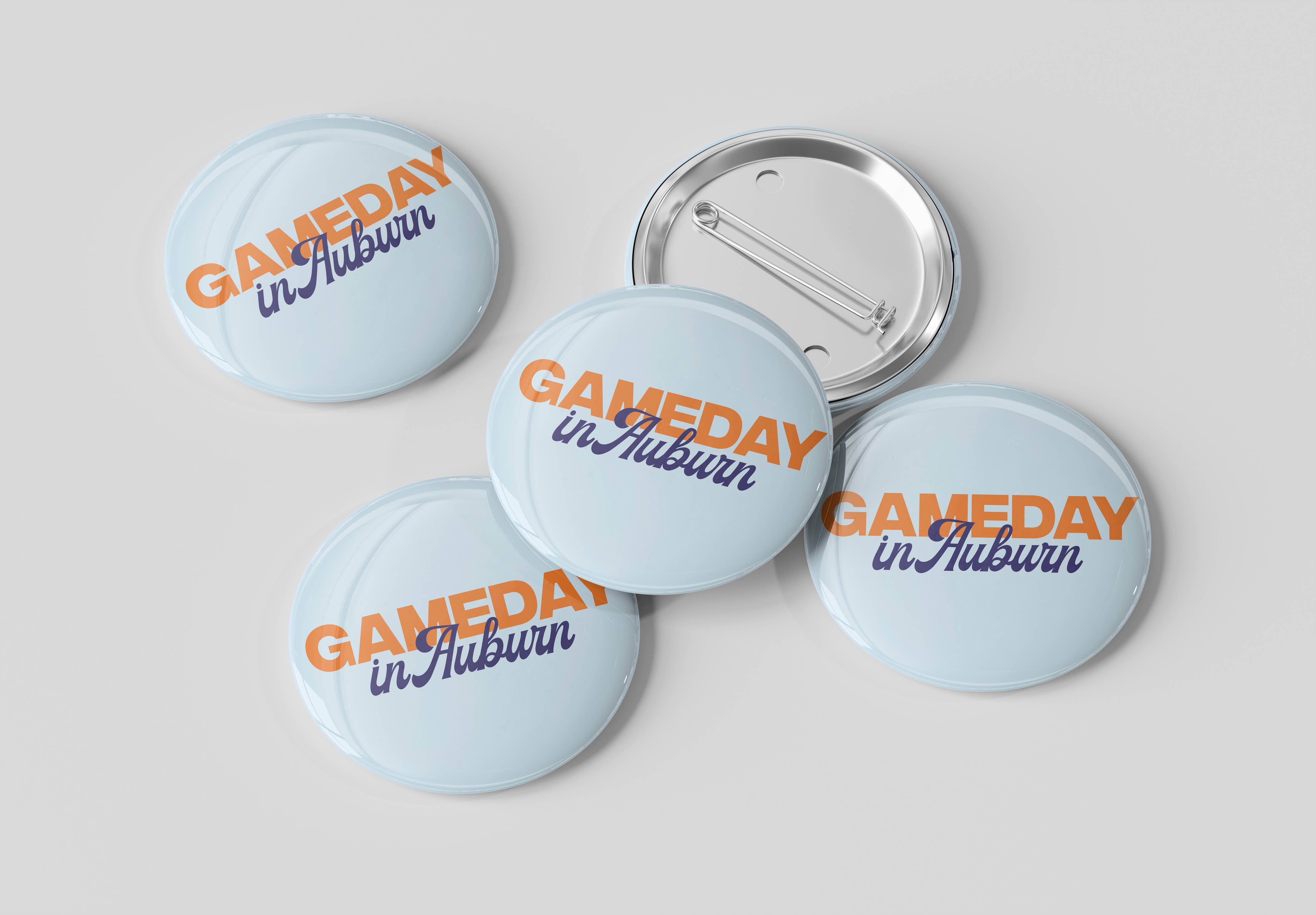 Five round pin-back buttons with a light blue background and the text 'GAMEDAY in Auburn' in orange and navy blue, with one button showing the silver metal pin-back mechanism, on a light gray background.