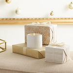 White candle on a beige surface with wrapped gifts and decorative lights in the background
