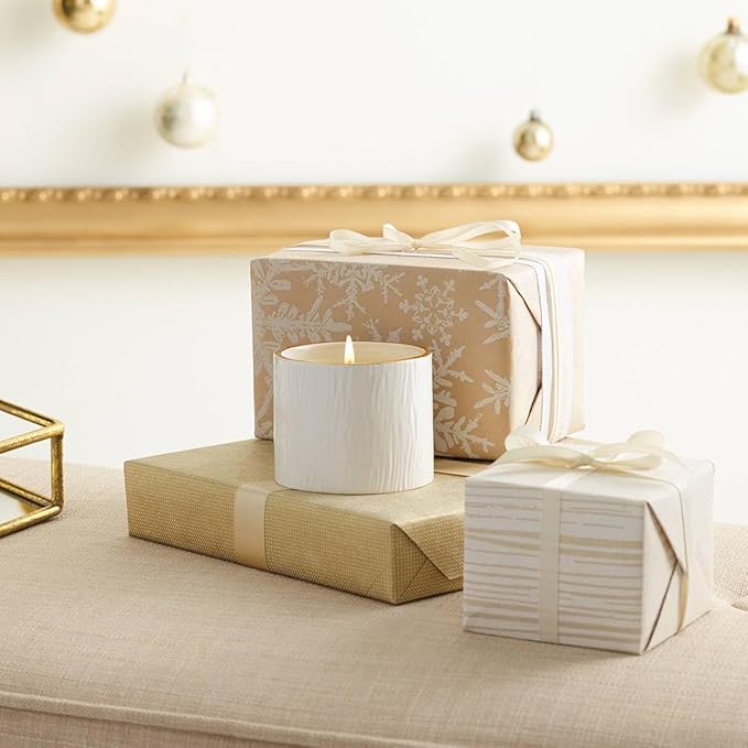 White candle on a beige surface with wrapped gifts and decorative lights in the background