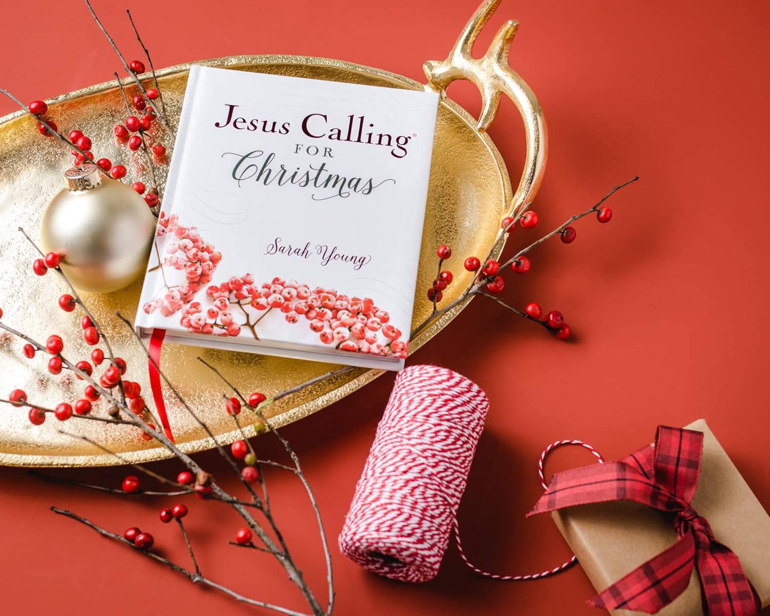 Book titled 'Jesus Calling for Christmas' by Sarah Young on a decorative tray with red berries and a gift, against a red background.
