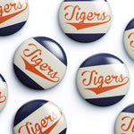 Several round glossy buttons with the word "Tigers" in orange script over diagonal navy and off-white stripes, arranged on a white background.