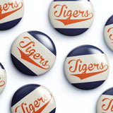 Several round glossy buttons with the word "Tigers" in orange script over diagonal navy and off-white stripes, arranged on a white background.