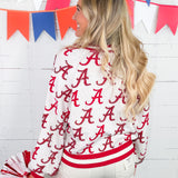Woman wearing a white jacket with red 'A' pattern, holding pom-poms, in front of colorful flags.