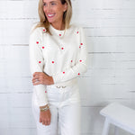 Woman wearing a white sweater with red heart patterns and white pants against a white wooden paneled wall.