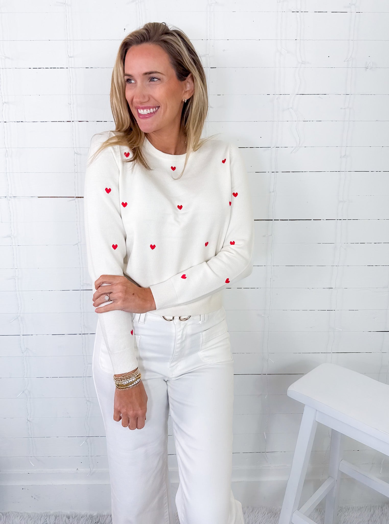 Woman wearing a white sweater with red heart patterns and white pants against a white wooden paneled wall.