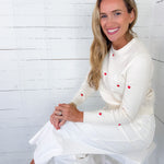 Woman wearing a white dress with red buttons sitting on a white stool against a white wooden wall.