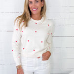 Woman wearing a white sweater with red heart patterns against a white wooden panel background