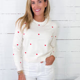 Woman wearing a white sweater with red heart patterns against a white wooden panel background