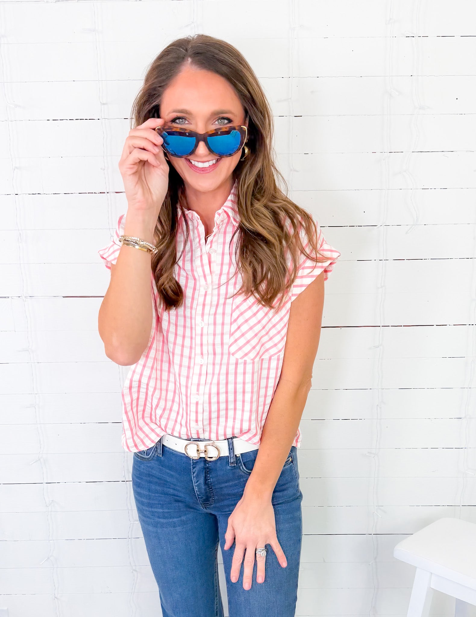 Woman wearing a red and white checkered shirt, blue jeans, and sunglasses against a white wooden background
