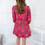 Person wearing a pink patterned dress standing against a white wooden panel wall.