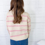 Person wearing a cream sweater with pink stripes and blue jeans against a white wooden panel background