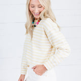 A woman with blonde hair wearing a cream and yellow striped long-sleeve top and white pants, standing against a white wood-paneled wall.