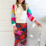 Woman wearing a colorful sweater and patterned skirt standing next to a clear chair against a white wooden panel background.