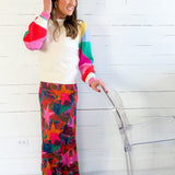 Woman wearing a colorful sweater and patterned pants standing against a white wooden panel wall.