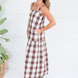 A woman modeling a brown and white gingham plaid midi dress with pockets, standing in front of a white wood-paneled wall next to a potted plant.