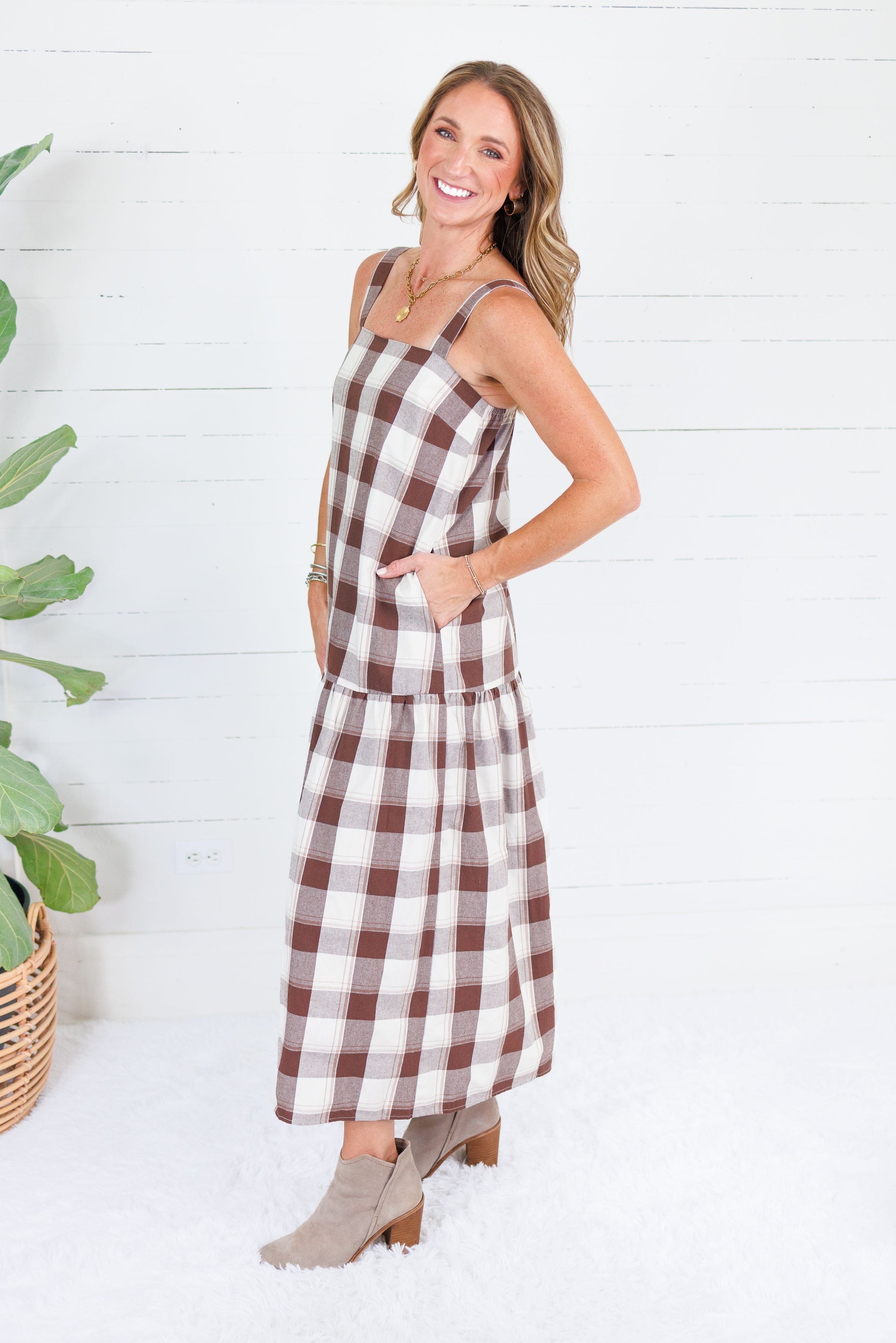 A woman modeling a brown and white gingham plaid midi dress with pockets, standing in front of a white wood-paneled wall next to a potted plant.