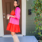 Woman in a pink and red dress with beige knee-high boots standing outside a house.