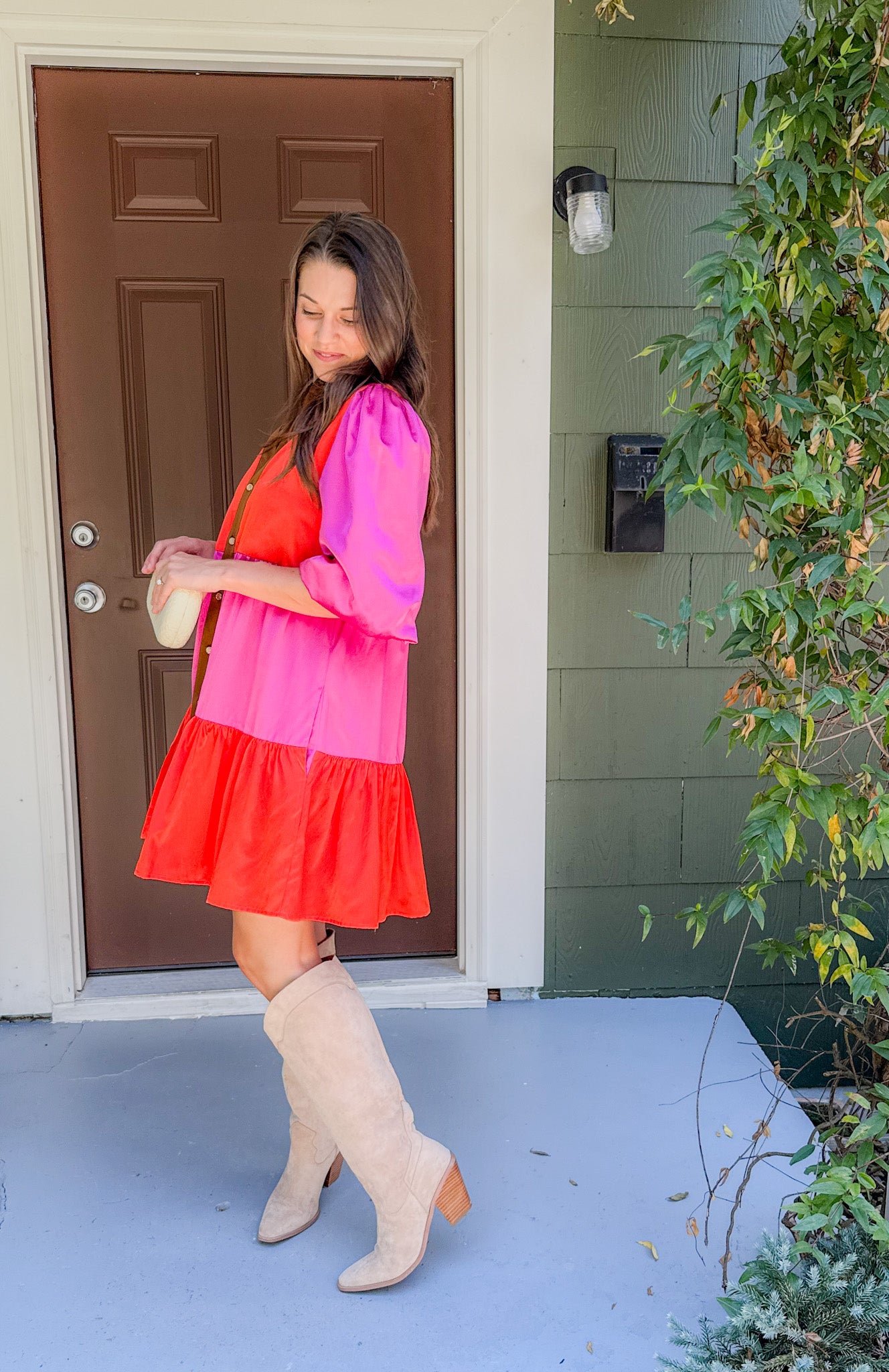 Woman in a pink and red dress with beige knee-high boots standing outside a house.