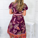Woman wearing a purple patterned dress sitting on a white chair against a white wooden panel background