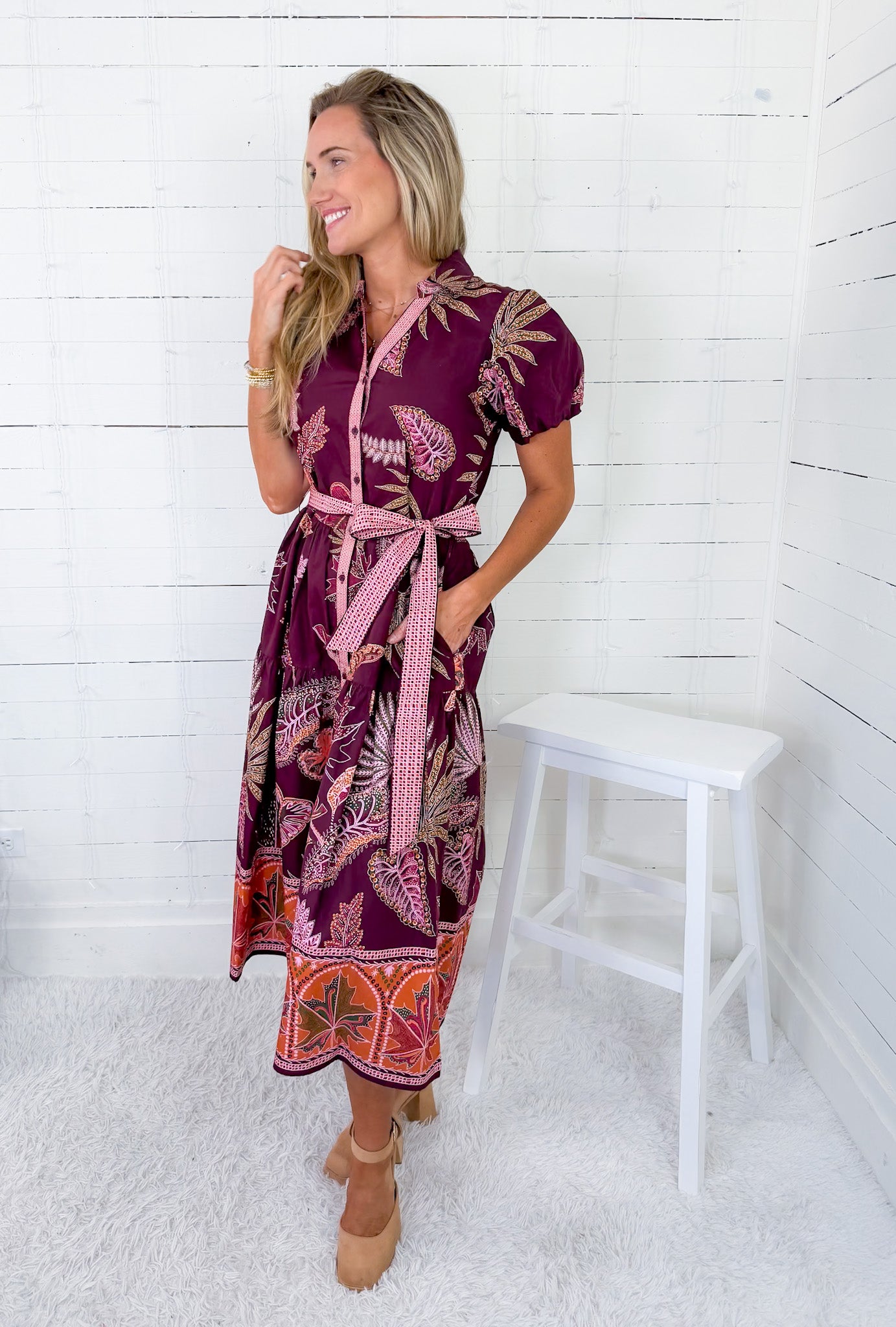 Woman wearing a patterned dress standing in a room with white walls and a white stool.