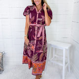 Woman wearing a patterned dress standing against a white wooden panel wall.
