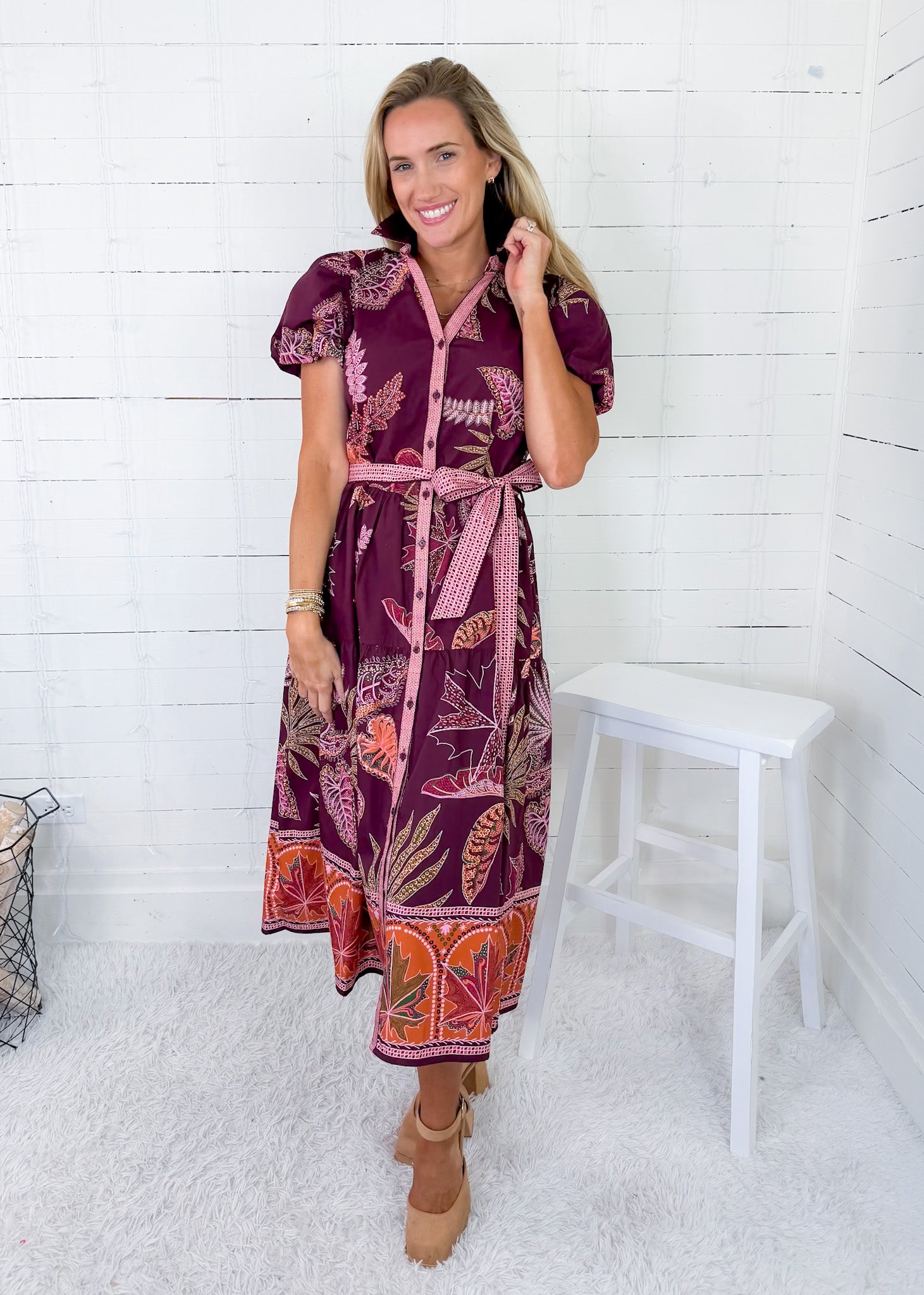 Woman wearing a patterned dress standing against a white wooden panel wall.
