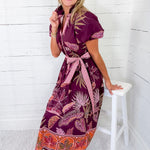 Woman wearing a patterned dress sitting on a white stool against a white wooden paneled wall.