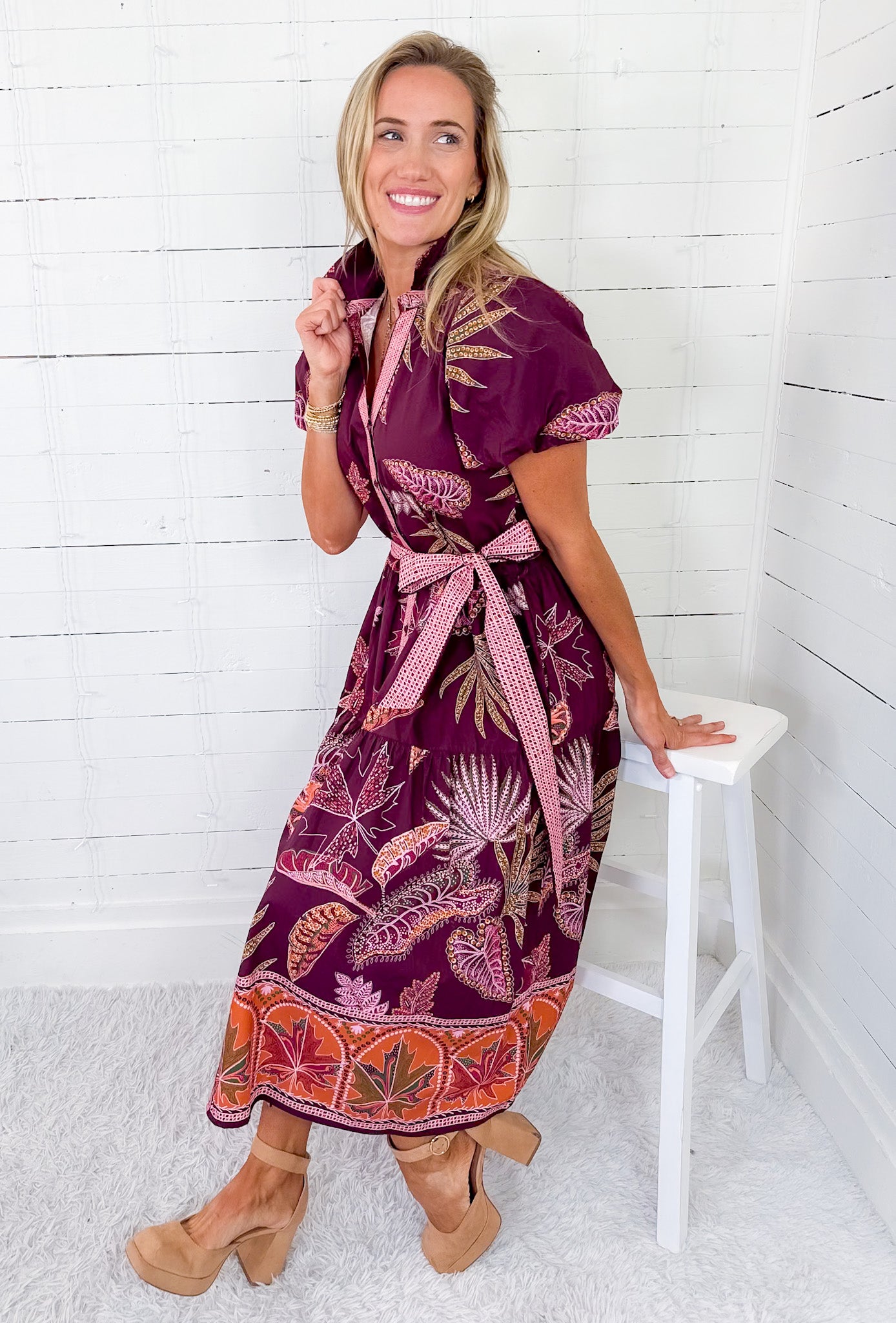 Woman wearing a patterned dress sitting on a white stool against a white wooden paneled wall.